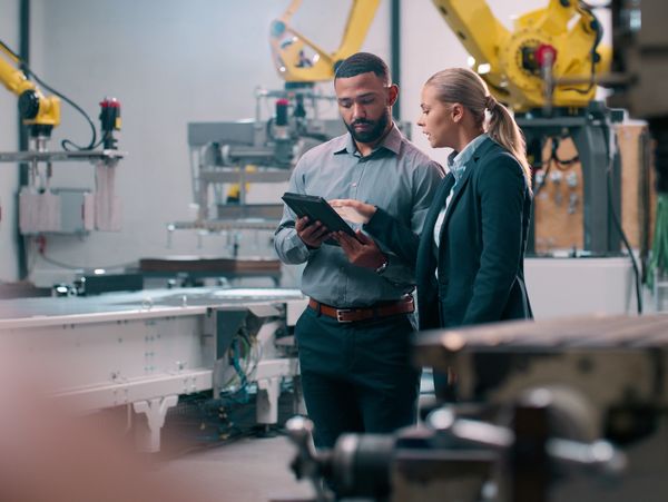 Two professionals discuss work on a tablet in a robotic manufacturing facility.