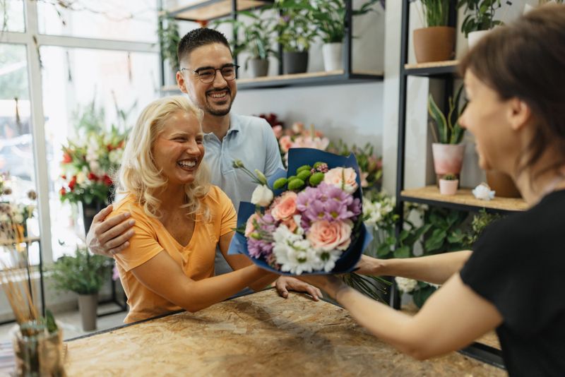 A happy couple receives a colorful bouquet from a florist at a flower shop, sharing smiles and joy in a moment filled with love, surrounded by a lush array of flowers and plants.
