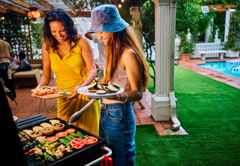 Two women preparing and grilling vegetables at an outdoor barbecue party. Social gathering with food. Summer cooking and celebration concept. Meat free shot.