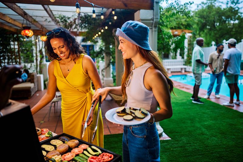 Two women preparing and grilling vegetables at an outdoor barbecue party. Social gathering with food. Summer cooking and celebration concept. Meat free shot.