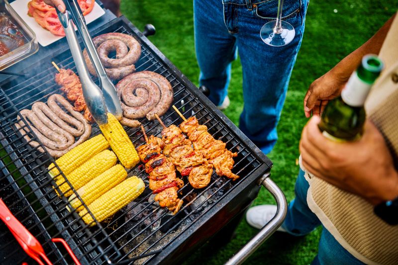 Close-up of hands grilling sausages, corn, and chicken skewers. Outdoor barbecue or Braai gathering. Summer food and cooking concept.