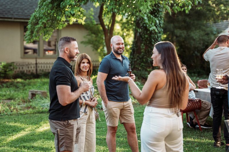 A small group of friends stand outside in a backyard, enjoying drinks and casual conversation during the wedding rehearsal dinner for their friends