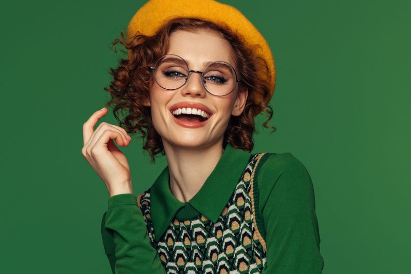 This vibrant portrait features a joyful young woman in a stylish retro outfit, highlighting a green blouse and a patterned vest. She wears a yellow beret and round glasses, complementing her curly red hair. The background is a solid green, enhancing the overall color harmony of the image. The woman's wide, happy smile and playful gesture of adjusting her glasses add a lively and cheerful vibe, making the photograph exude positive energy and vintage charm.
