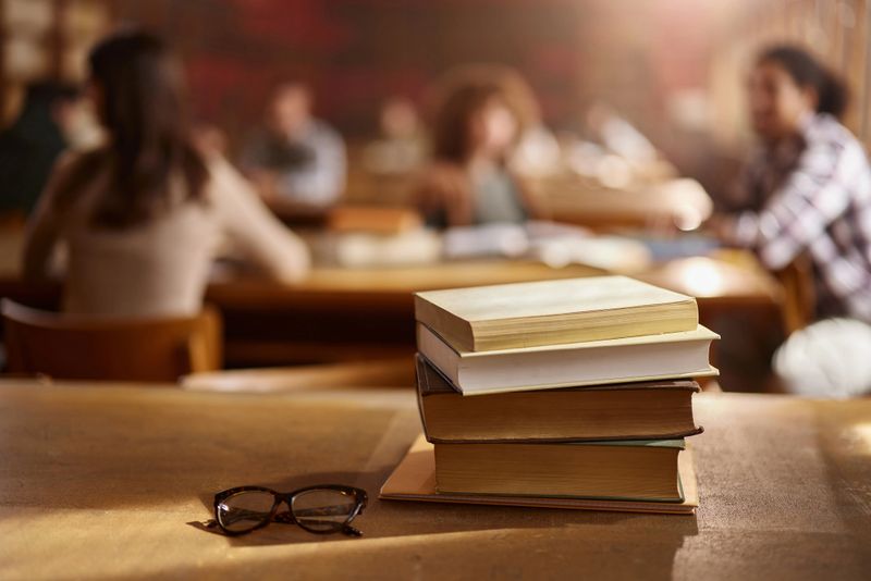Close up of pile of books on a desk in library with people in the background.
