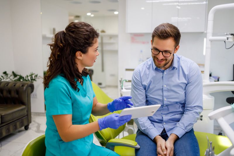 A dentist in blue scrubs and gloves is holding a tablet and discussing treatment options with a male patient in a dental clinic with modern furnishings and equipment.