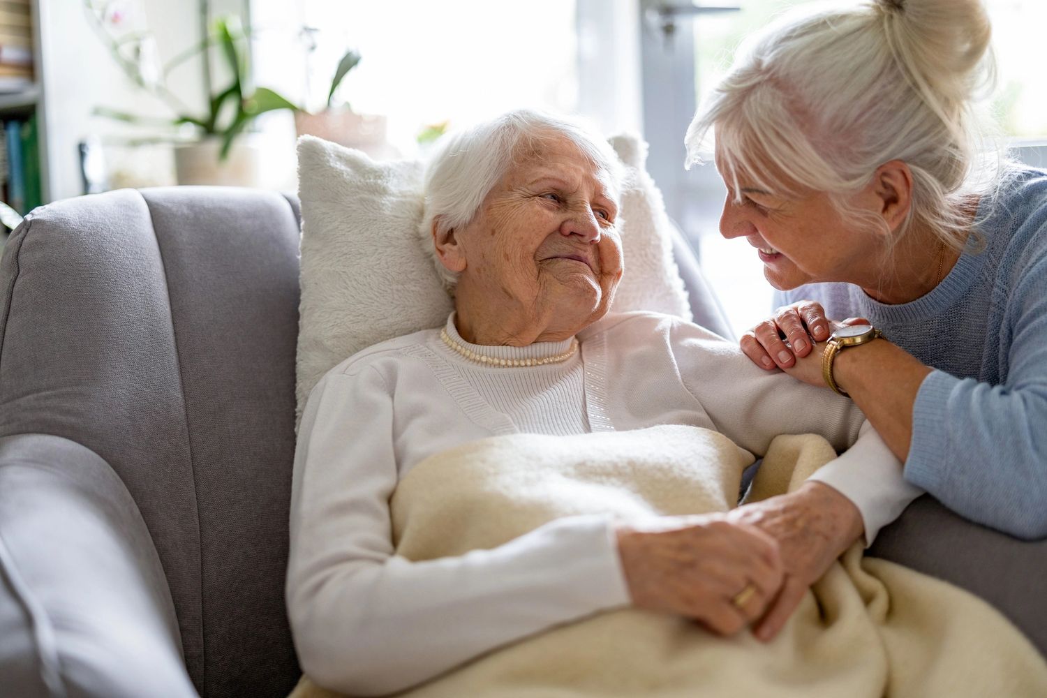 Elderly woman resting on a couch with a smiling visitor holding her hand.