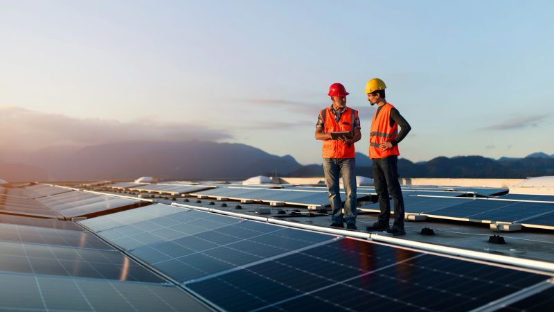 Two technicians in reflective vests checking solar power plant installed on industrial building roof. Alternative energy source, ecology and carbon footprint reduction concept.