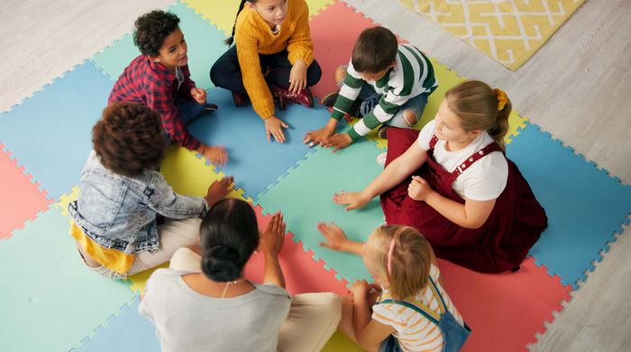Children and a teacher sitting in a circle playing a hand game on colorful foam mats.