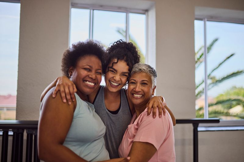 Confident young female coach hugging her multiethnic students, looking and smiling cheerfully together at camera after class in studio