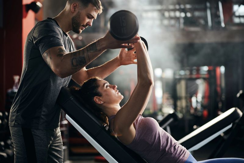 Young man helping his girlfriend during her sports training with barbell in a health club.