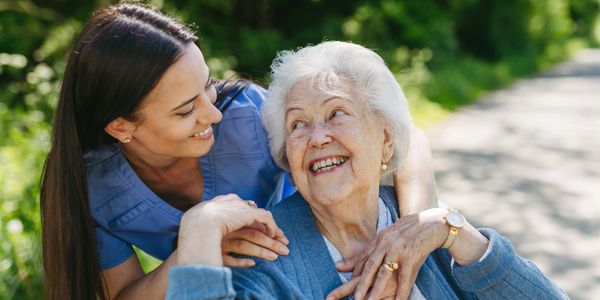 A caregiver warmly embraces an elderly woman outdoors, both smiling joyfully.