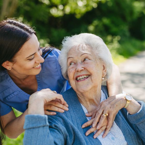 A caregiver warmly embraces an elderly woman outdoors, both smiling joyfully.