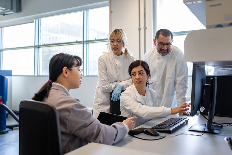 Medium shot of scientists recording research on a computer. They are all looking at the screen while wearing protective lab coats.Videos are available similar to this scenario.