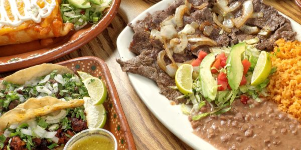 Mexican meal with tacos, grilled steak, rice, beans, and fresh garnish.
