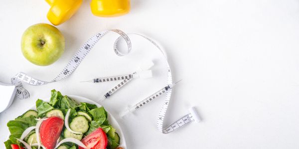 Healthy salad, green apple, measuring tape, and yellow dumbbells arranged on a white surface.