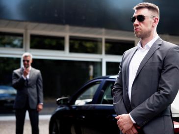 Two men in suits standing near a black car, one wearing sunglasses.
