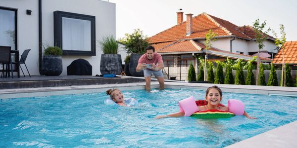 Two children playing in a pool with a man watching nearby on a sunny day.