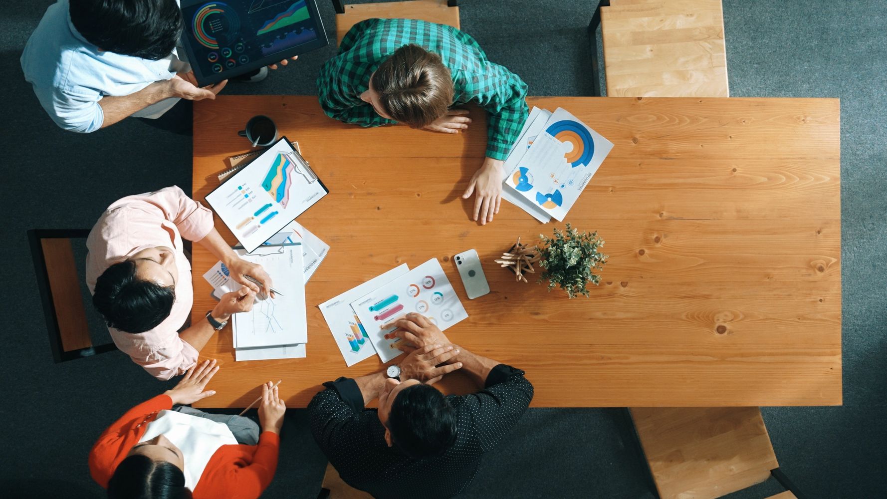 Top view of five people analyzing charts and data around a wooden table.