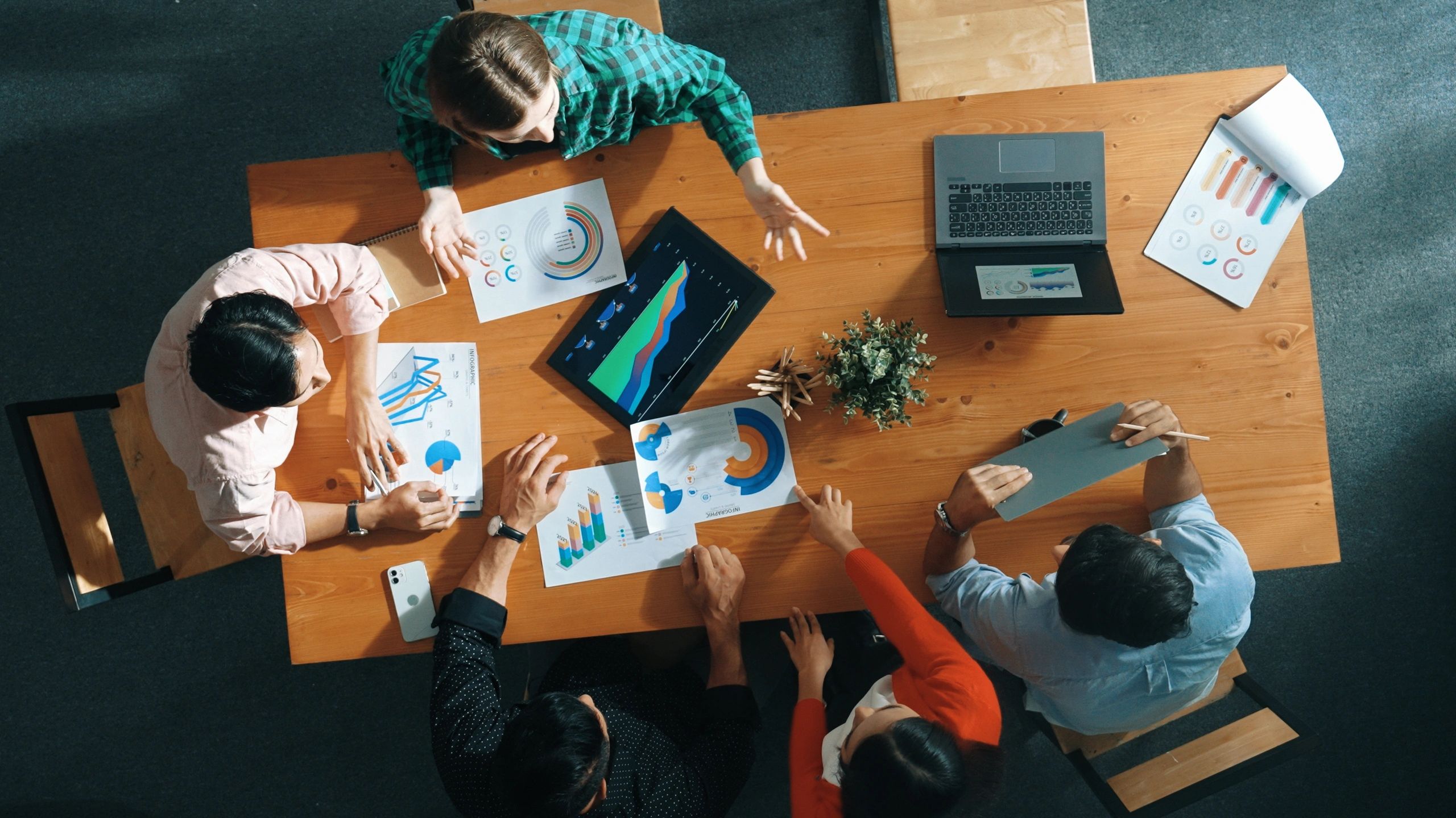 A team collaborates over charts and laptops at a wooden table in a meeting room.