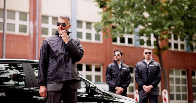 Luxury car business security guard providing VIP protection services, equipped with walkie-talkie, safeguarding high-end client outside corporate event.