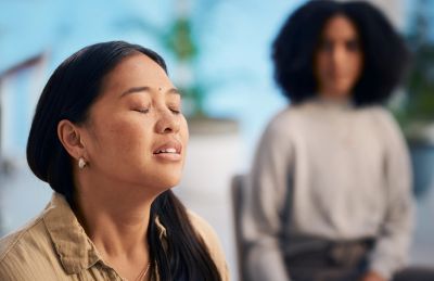 A woman peacefully meditates with eyes closed, another person blurred in the background.