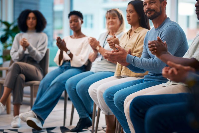A diverse group of people sitting in a circle and clapping hands during a support group meeting. This image captures the sense of community, support, and connection among individuals from different backgrounds.