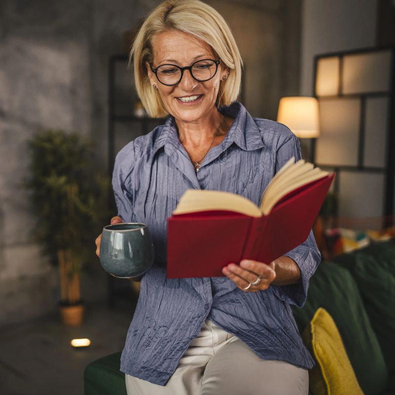 Mature senior woman sit in the living room read a book and drink coffee