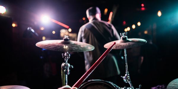 Drummer playing on stage with a singer in the background under colorful lights.