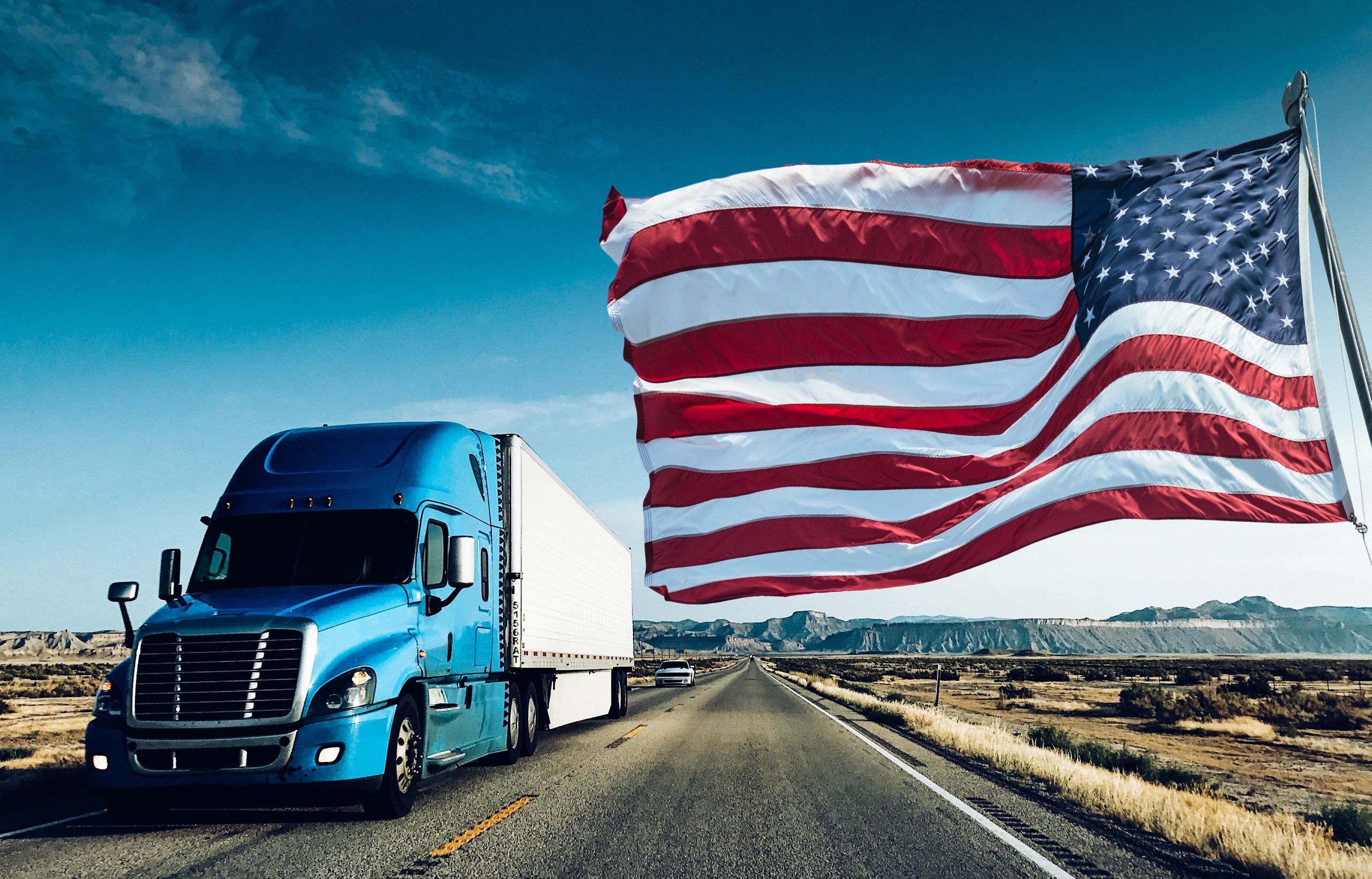 A blue semi-truck on a highway with a large American flag waving.