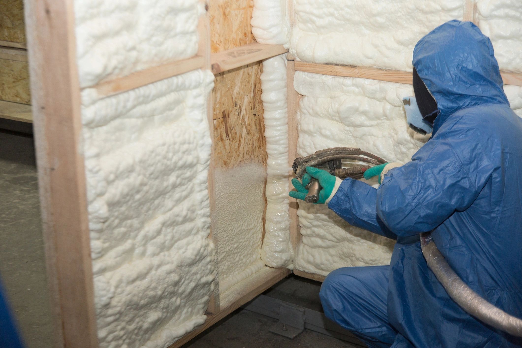 Person spraying foam insulation on wall panels wearing protective gear.