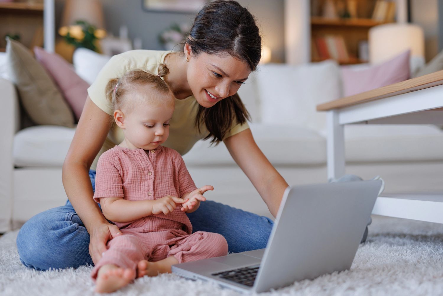 A mother and toddler using a laptop together on a carpeted floor.
