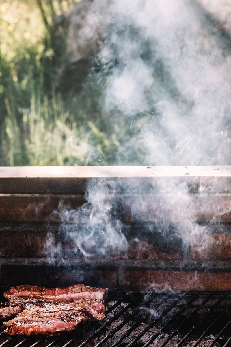 A close-up shot of grilled meat on a brick barbecue. The meat is sizzling and smoking, with white smoke billowing up from the grill