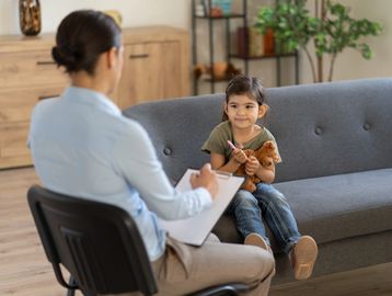 A young girl holds a teddy bear during a therapy session with a professional.