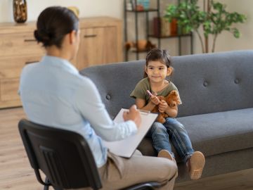 A young girl holds a teddy bear during a therapy session with a professional.