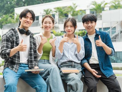 Four young adults sitting outdoors, smiling, and giving thumbs up.