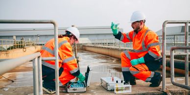Two workers in orange safety gear testing water samples at an industrial site.