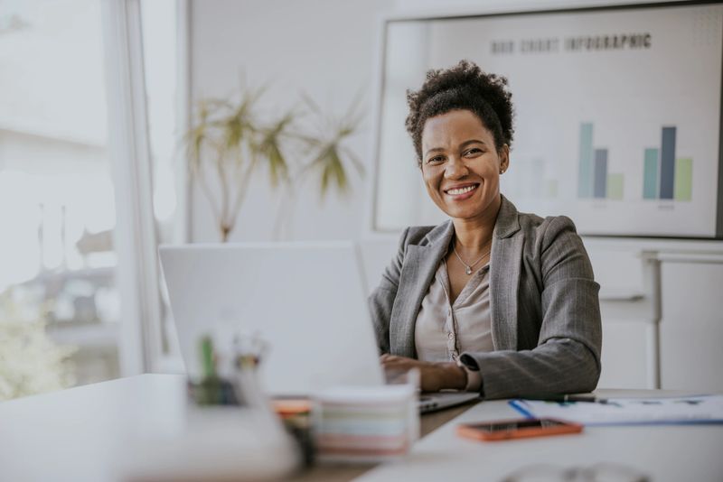 A smiling businesswoman sits at a desk in a modern office, working on a laptop. There is a window behind her, and a whiteboard with charts is visible in the background. She is wearing a blazer and has a necklace on. The desk is clean and organized.
