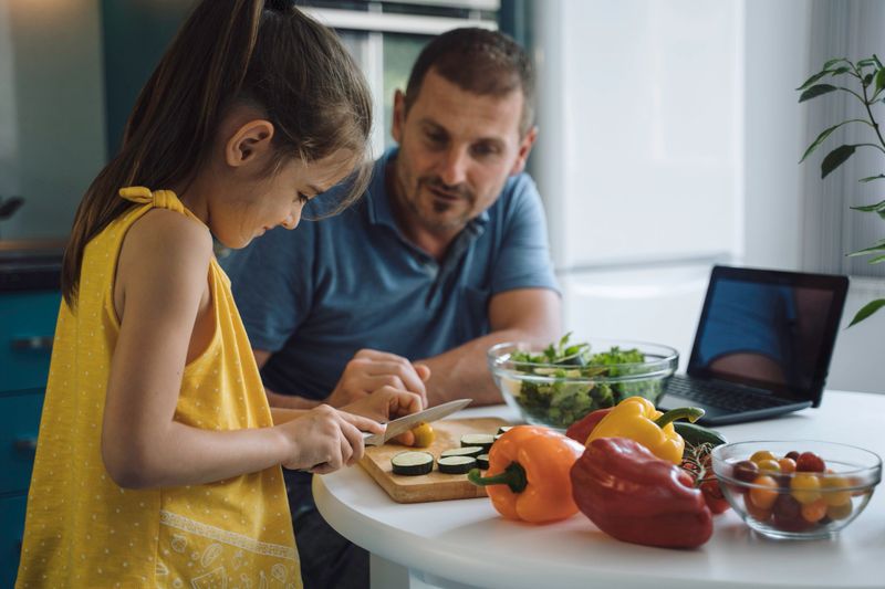 A father supervises his young daughter chopping vegetables in a modern kitchen, showcasing the principles of guidance, learning, and family involvement in cooking.