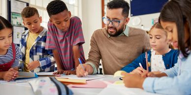 Teacher helping young students with writing at a classroom table.