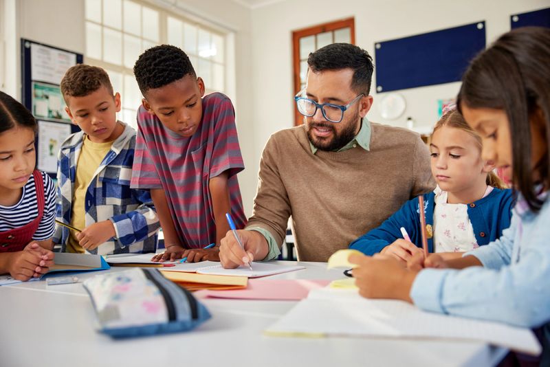 Young school teacher helping elementary students while writing in notebooks. Hispanic male teacher wearing spectacles and checking student notebooks after test while talking to them in classroom. Happy male teacher helping and checking works of pupil in classroom.
