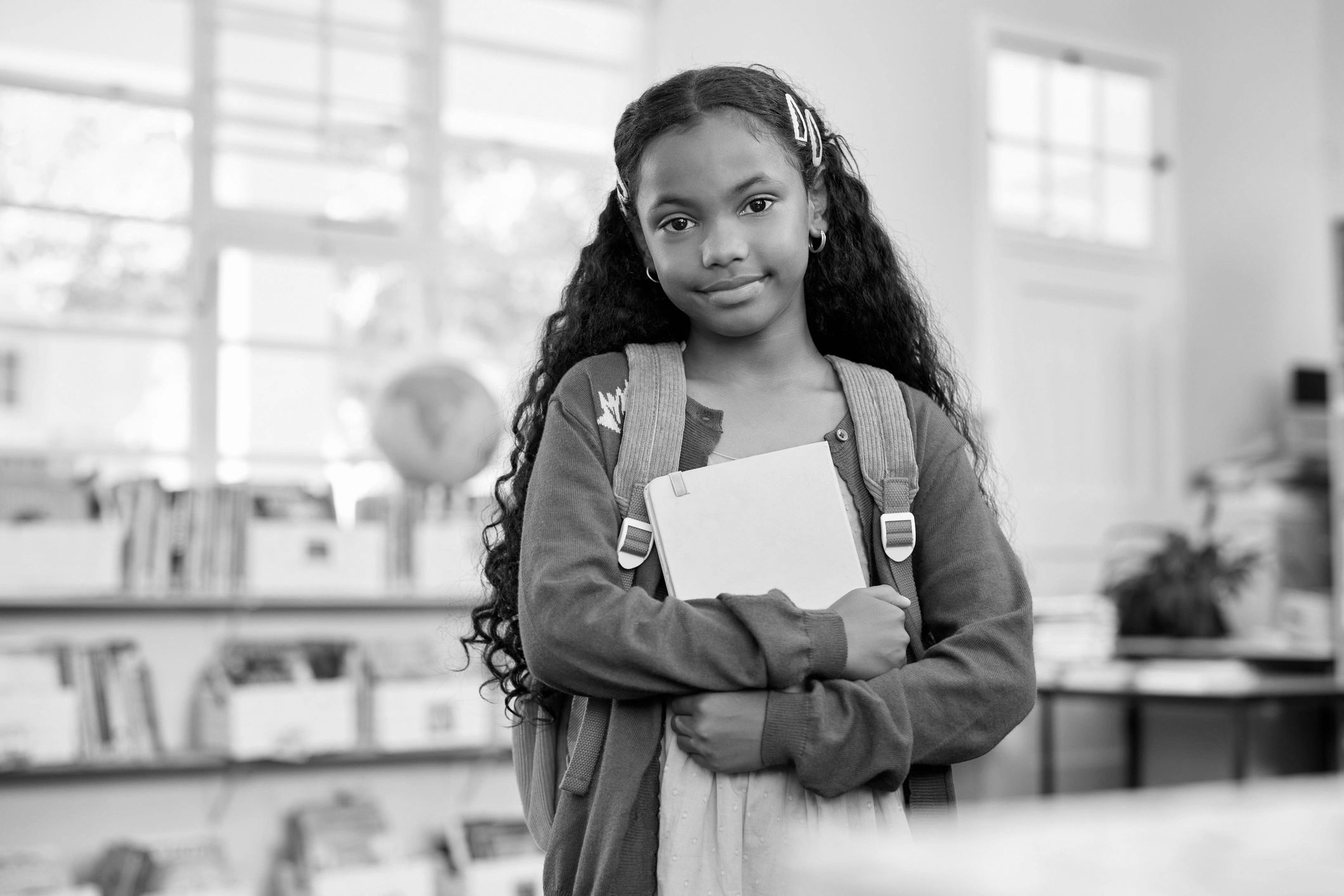 Young girl with backpack holding a book in a classroom.