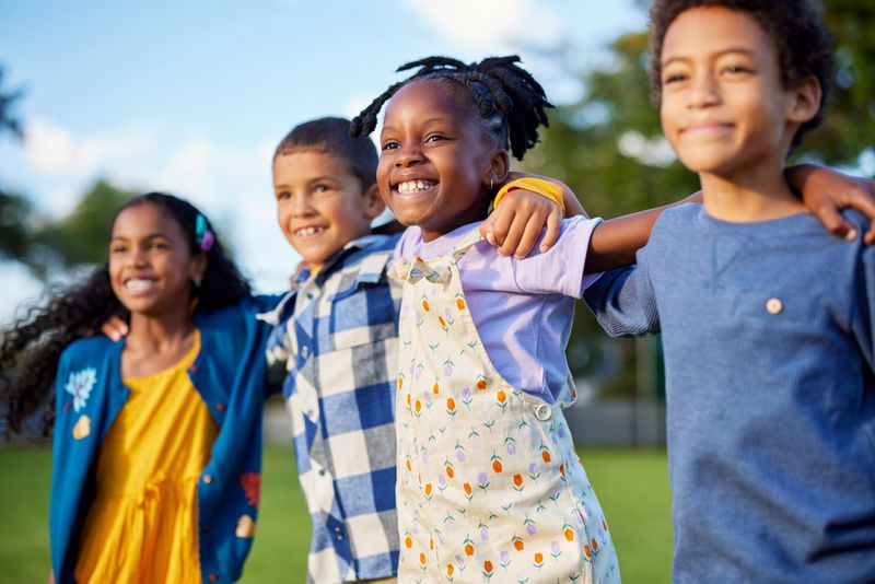 Side view of little children jumping together in a row while looking their bright future. Happy little boys and smiling cute girls making friends and having fun together at park. Cheerful multiethnic kids enjoying summertime and embracing in outdoor park at sunset during summer camp.