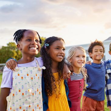 Five diverse children smiling with arms around each other outdoors at sunset.