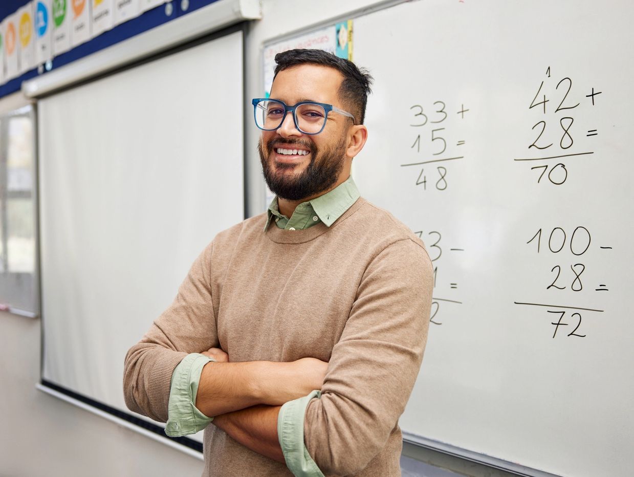 Smiling male teacher standing with arms crossed in front of a whiteboard with math problems.