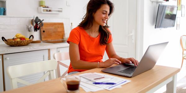 A woman in an orange shirt working on a laptop at a kitchen table.
