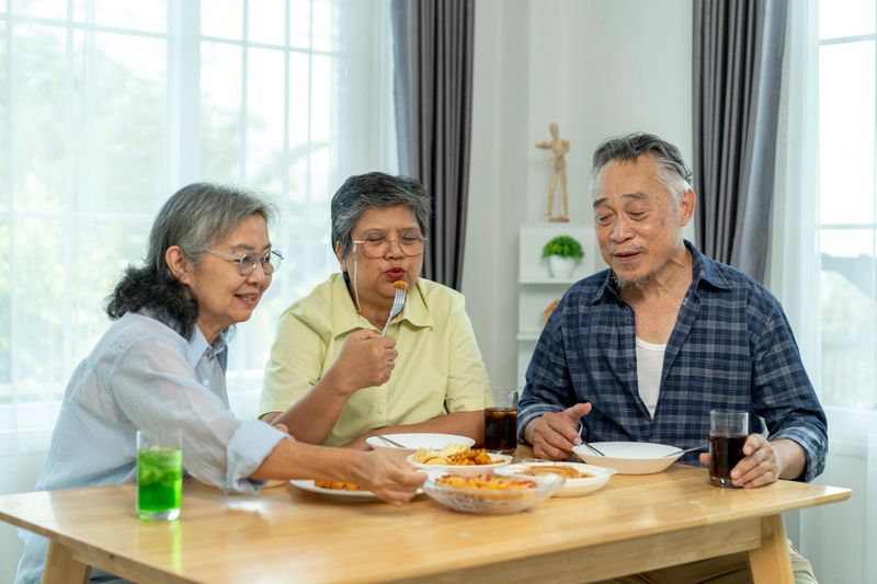 Three elderly Asian people enjoying a meal together at a table by a large window. They appear engaged in conversation and laughter while sharing food from plates on the table.