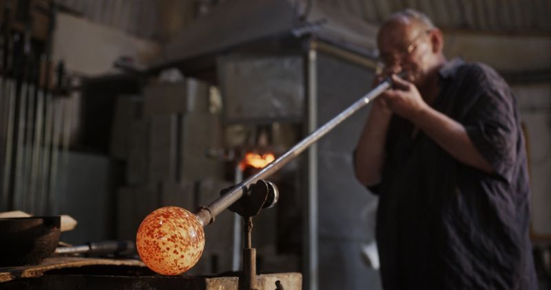 A master glassblower is seen inflating a vibrant molten glass vase, using a blowpipe to shape the glass into its final form in a traditional glassblowing workshop, with the furnace aglow in the background.