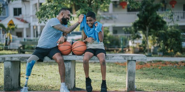 A man with a prosthetic leg and a boy share a joyful moment on a bench with basketballs.