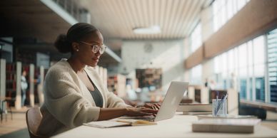 Woman in glasses working on a laptop in a bright library.