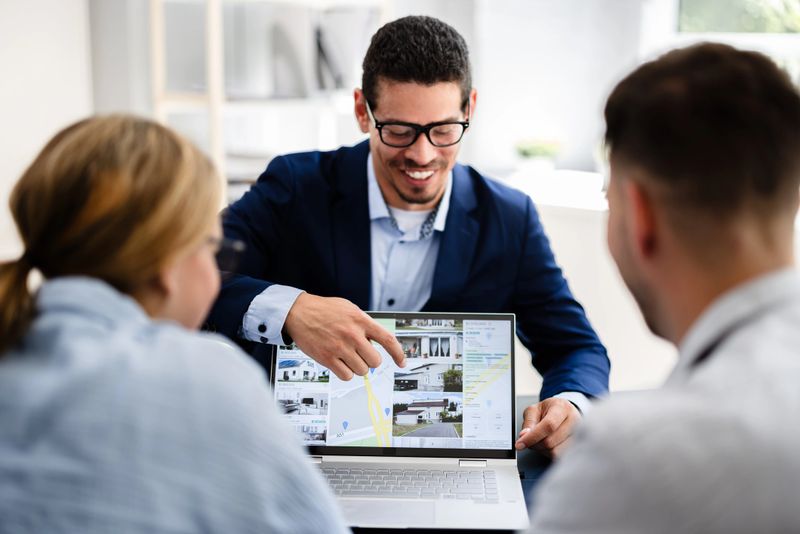 Professional Real Estate Agent Presenting Property Options to Happy Couple Using Laptop in Modern Office Setting